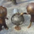 Two bronze child statues lean toward a rusted metal sphere sculpture in a plaza with a tree and cobblestone ground.