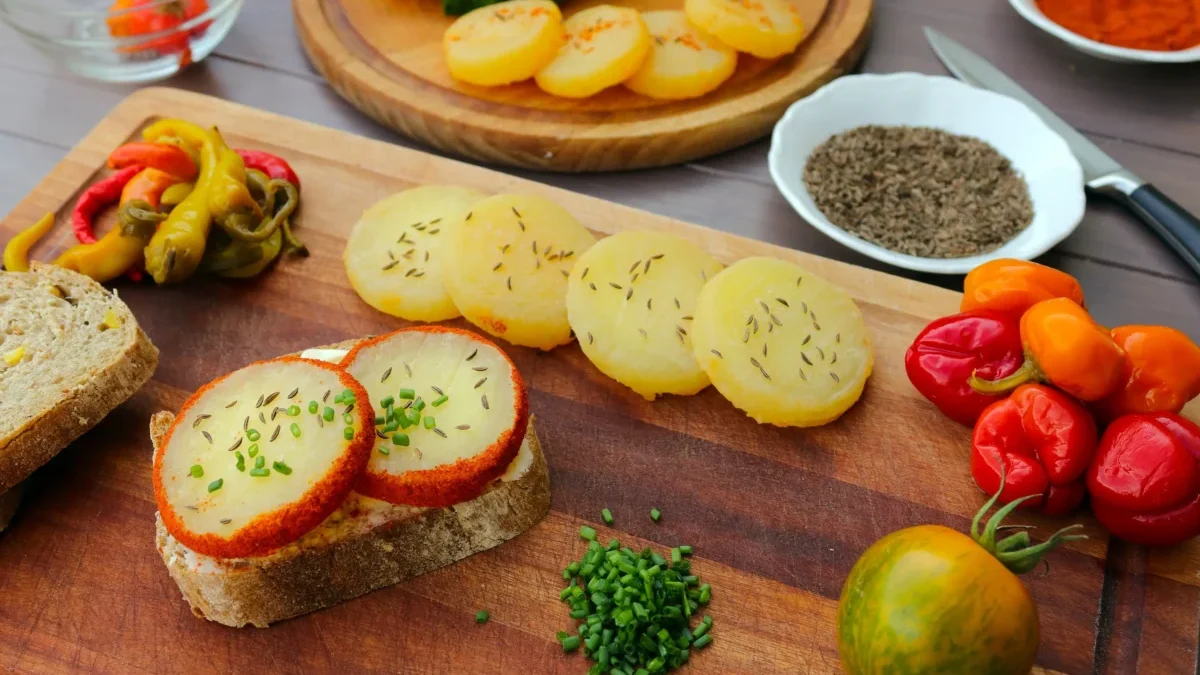 Assorted appetizers on a wooden board: potato rounds with herbs, sliced bread with herb cheese, and colorful peppers alongside cherry tomatoes.