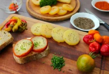 Assorted appetizers on a wooden board: potato rounds with herbs, sliced bread with herb cheese, and colorful peppers alongside cherry tomatoes.