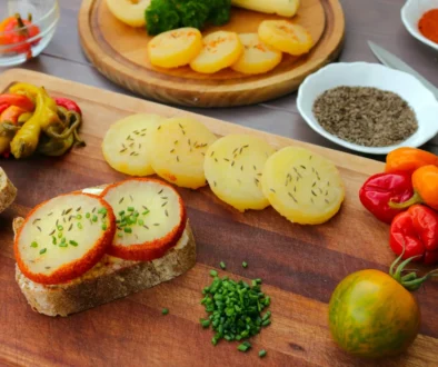 Assorted appetizers on a wooden board: potato rounds with herbs, sliced bread with herb cheese, and colorful peppers alongside cherry tomatoes.