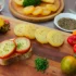 Assorted appetizers on a wooden board: potato rounds with herbs, sliced bread with herb cheese, and colorful peppers alongside cherry tomatoes.