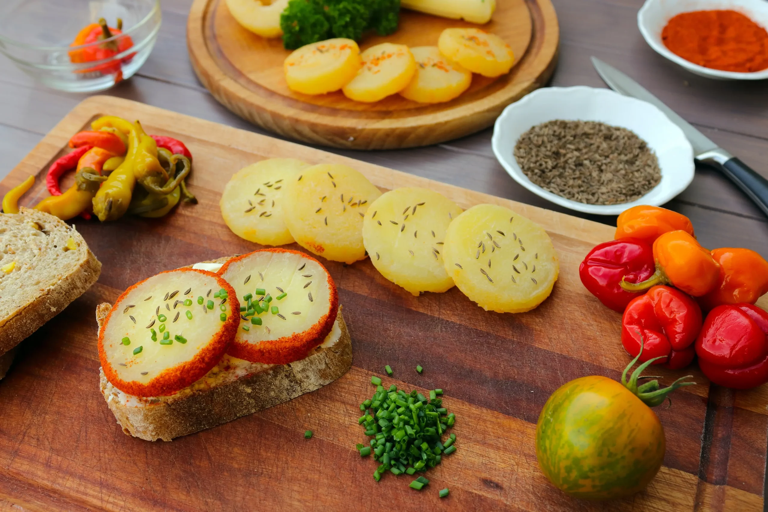 Assorted appetizers on a wooden board: potato rounds with herbs, sliced bread with herb cheese, and colorful peppers alongside cherry tomatoes.