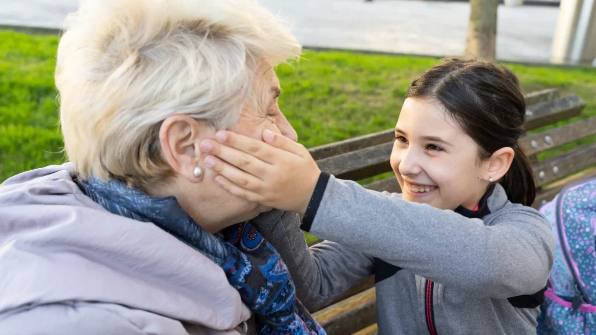 Young girl and an elderly woman laughing together on a park bench; the girl gently cups the older woman's cheek in a playful moment outdoors.