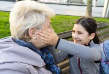 Young girl and an elderly woman laughing together on a park bench; the girl gently cups the older woman's cheek in a playful moment outdoors.