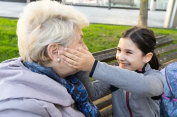 Young girl and an elderly woman laughing together on a park bench; the girl gently cups the older woman's cheek in a playful moment outdoors.