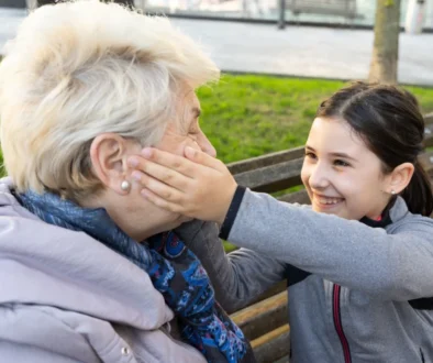 Young girl and an elderly woman laughing together on a park bench; the girl gently cups the older woman's cheek in a playful moment outdoors.