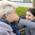 Young girl and an elderly woman laughing together on a park bench; the girl gently cups the older woman's cheek in a playful moment outdoors.