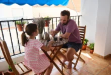Man and young girl sitting at a wooden patio table on a balcony, sharing a drink or snack outdoors.