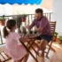 Man and young girl sitting at a wooden patio table on a balcony, sharing a drink or snack outdoors.