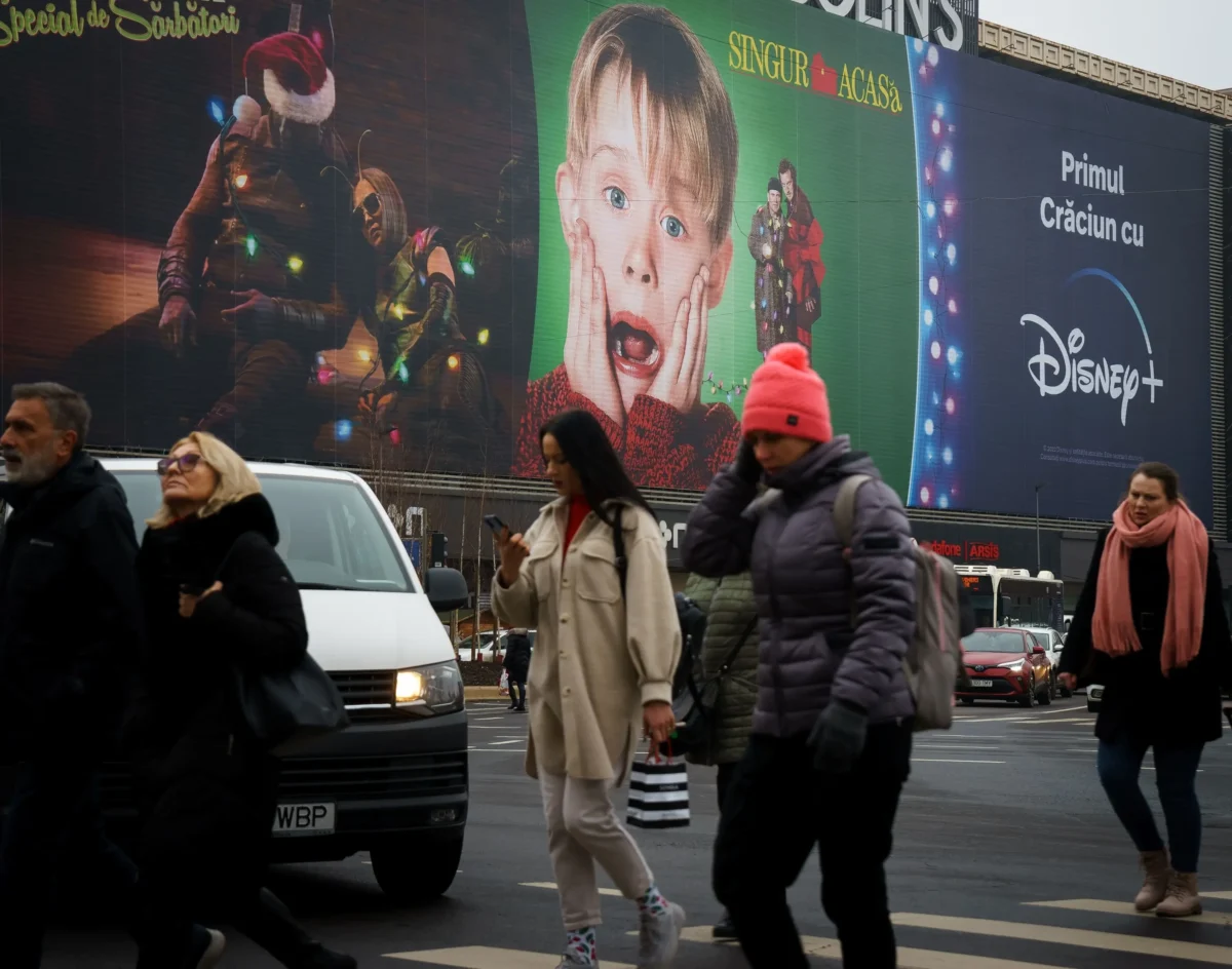 Kevin1 | TVGURU.cz Rušná městská ulice před velkým billboardem Disney+, chodci procházející kolem barevných vánočních reklam a vlevo viditelná čepice Santy. Uprostřed plakán Sám doma.