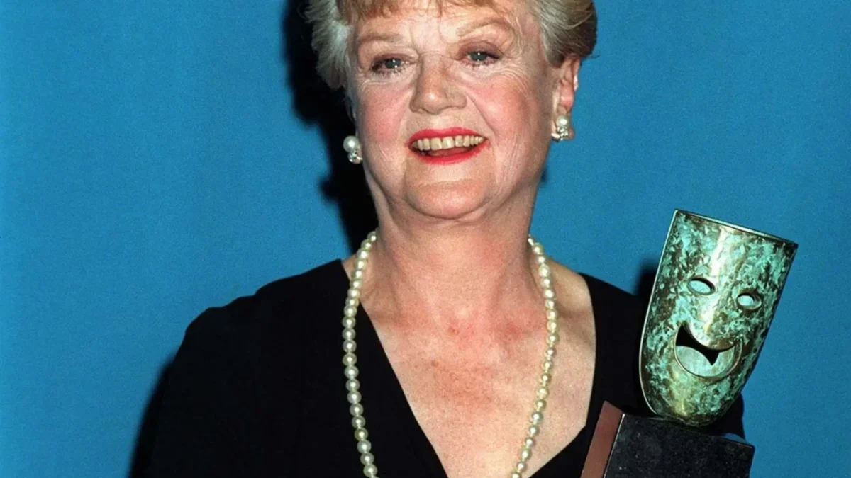 Older woman in a black dress with a pearl necklace, smiling, holding a gleaming award against a blue backdrop.