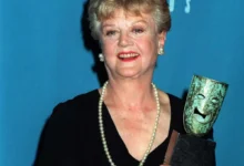 Older woman in a black dress with a pearl necklace, smiling, holding a gleaming award against a blue backdrop.