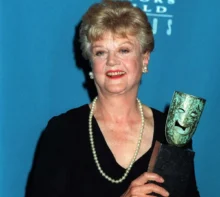 Older woman in a black dress with a pearl necklace, smiling, holding a gleaming award against a blue backdrop.