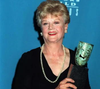 Older woman in a black dress with a pearl necklace, smiling, holding a gleaming award against a blue backdrop.