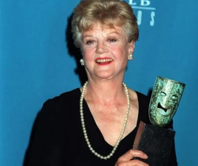 Older woman in a black dress with a pearl necklace, smiling, holding a gleaming award against a blue backdrop.