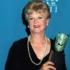 Older woman in a black dress with a pearl necklace, smiling, holding a gleaming award against a blue backdrop.