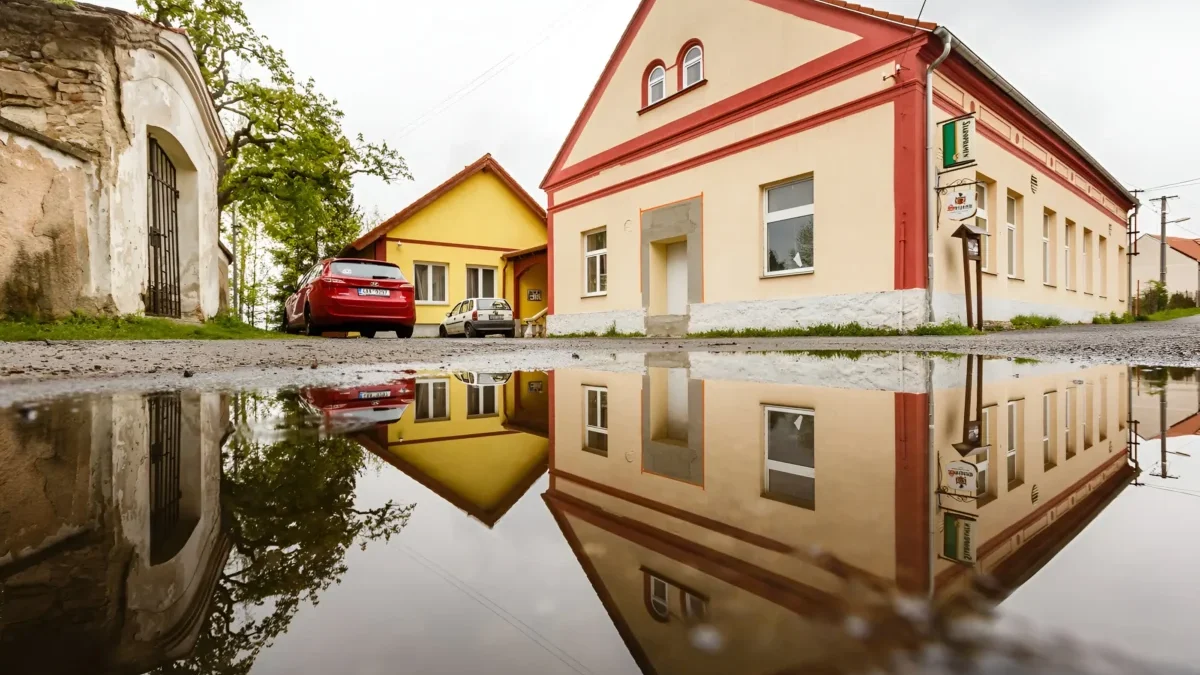 Street with pastel-colored buildings (peach, yellow) and red trim, reflected in a large puddle on the road.