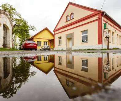 Street with pastel-colored buildings (peach, yellow) and red trim, reflected in a large puddle on the road.