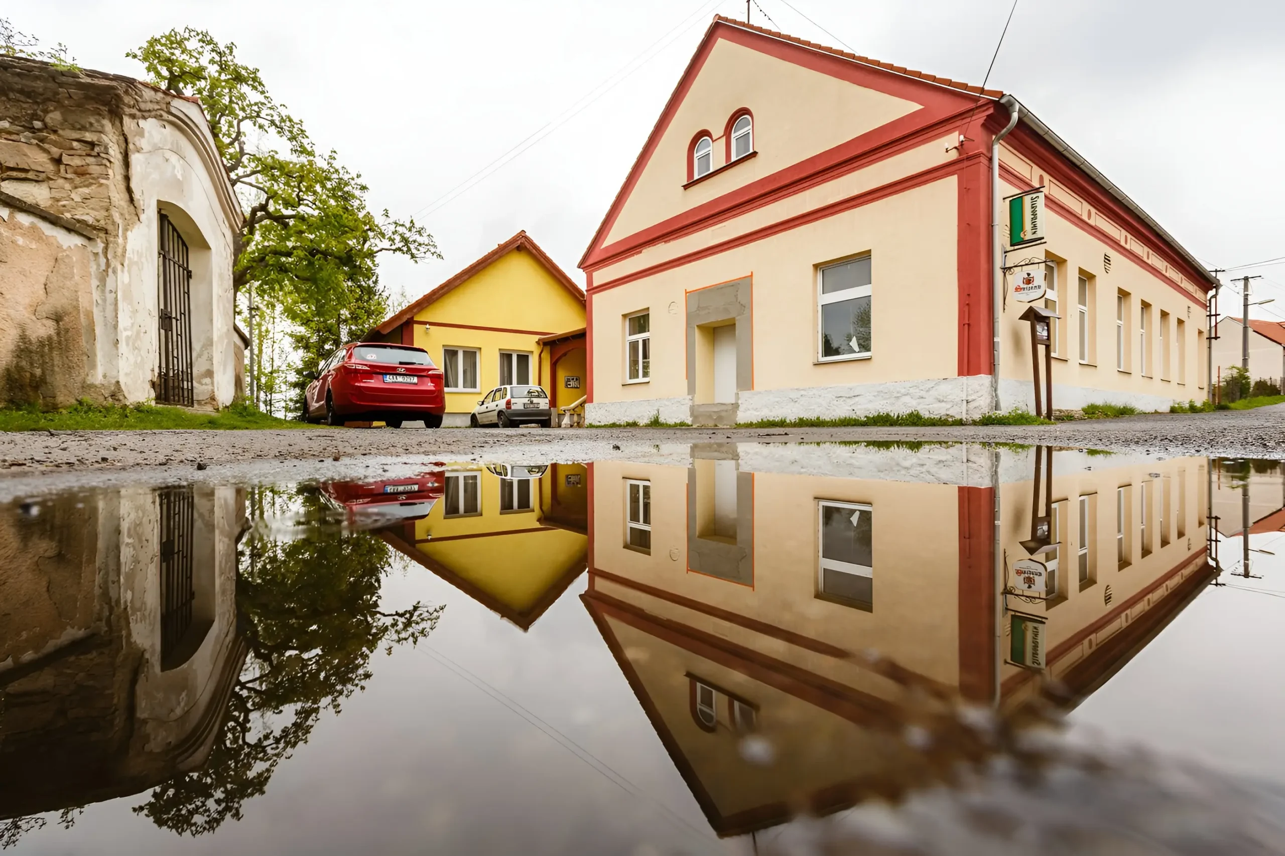 Street with pastel-colored buildings (peach, yellow) and red trim, reflected in a large puddle on the road.