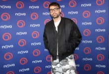 Man with short dark hair and glasses posing in a black bomber jacket over a white shirt and gray camouflage pants in front of a blue Nova backdrop with orange circular logos.