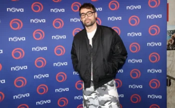 Man with short dark hair and glasses posing in a black bomber jacket over a white shirt and gray camouflage pants in front of a blue Nova backdrop with orange circular logos.