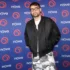 Man with short dark hair and glasses posing in a black bomber jacket over a white shirt and gray camouflage pants in front of a blue Nova backdrop with orange circular logos.