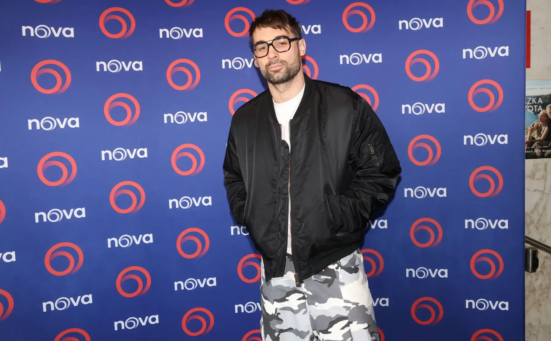 Man with short dark hair and glasses posing in a black bomber jacket over a white shirt and gray camouflage pants in front of a blue Nova backdrop with orange circular logos.