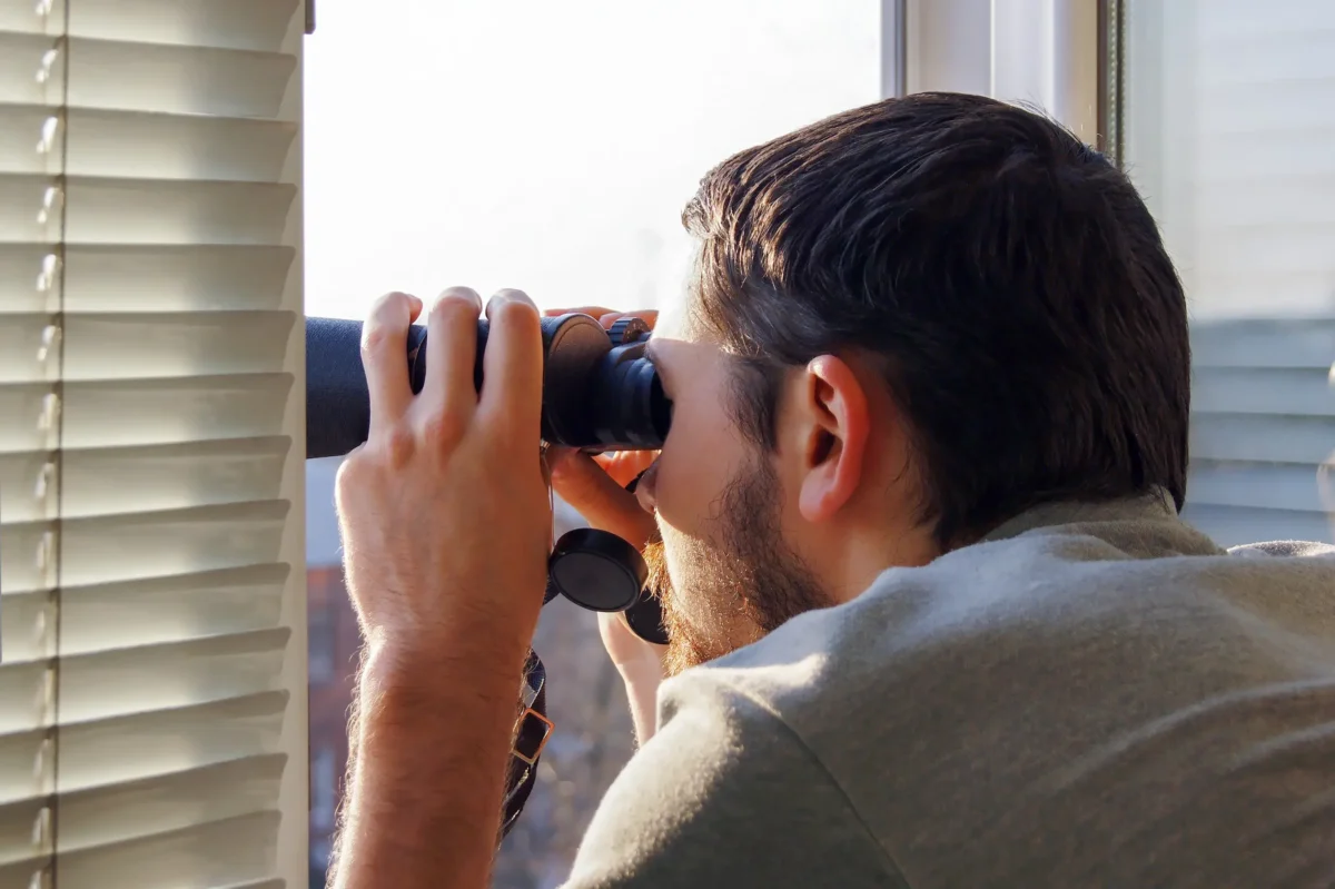 Hlavní postava si zpestřuje život šmírováním sousedů. Zdroj: Shutterstock | TVGURU.cz Muž dívající se z okna dalekohledem