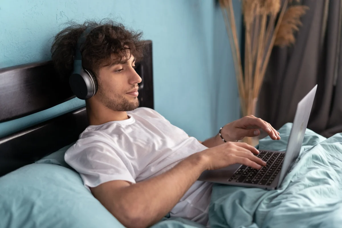shutterstock_2325770113 1 | TVGURU.cz Young man lying in bed with over-ear headphones on, using a laptop on his lap
