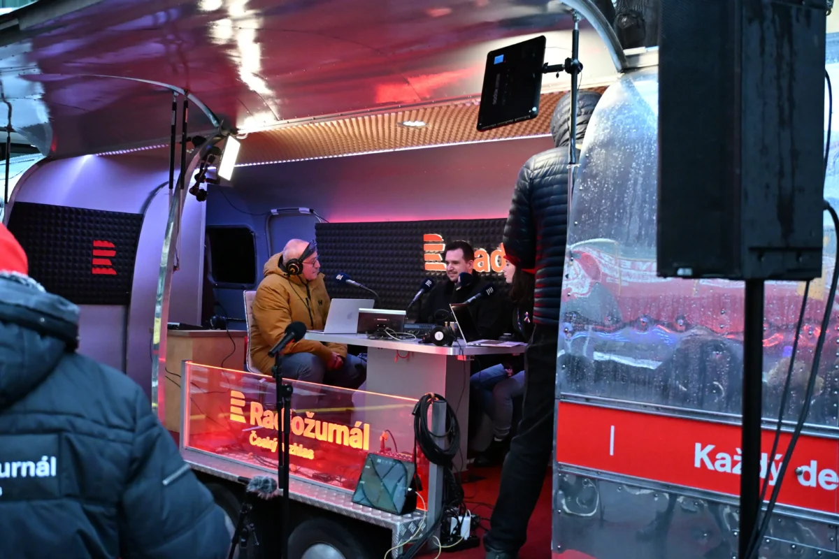 shutterstock_2703347835 1 | TVGURU.cz Three people sit at a desk inside a mobile radio studio, wearing headphones and speaking into microphones with laptops and broadcast gear around them.