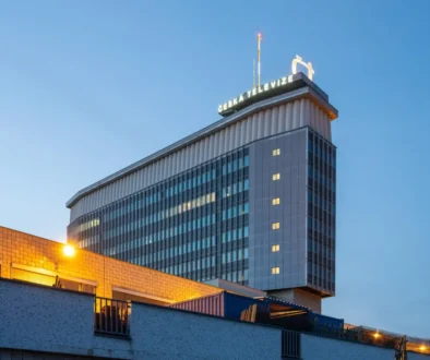 Tall glass-clad office tower with a lit Česká Televize sign on the rooftop, photographed at dusk against a blue sky.