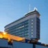 Tall glass-clad office tower with a lit Česká Televize sign on the rooftop, photographed at dusk against a blue sky.