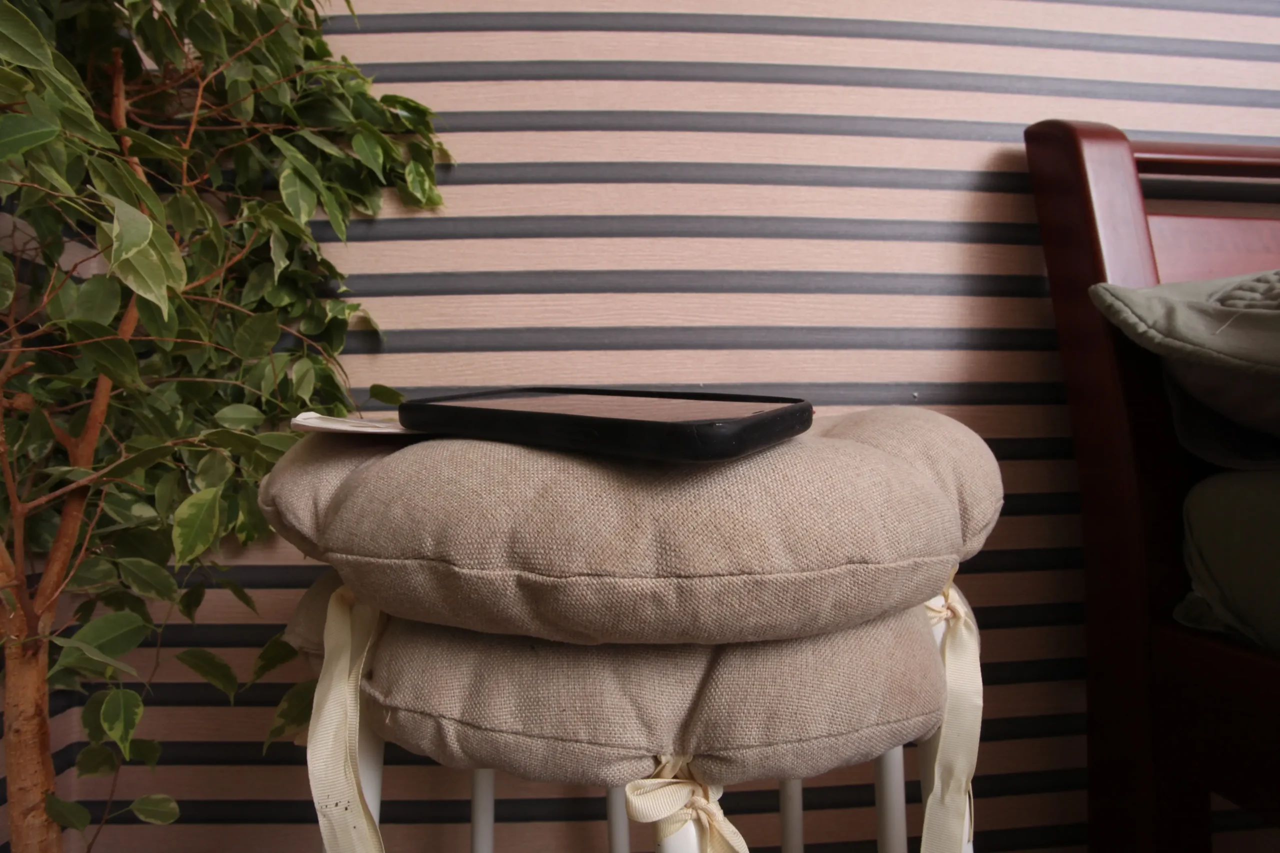 Beige cushioned stack with a black tablet resting on top, a leafy plant on the left, and a wooden-framed chair on the right against striped walls.