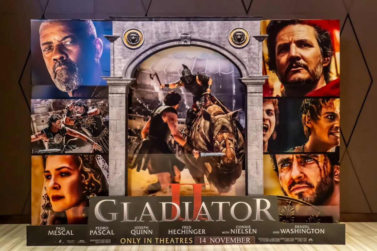 gladiator2 | TVGURU.cz Kolážový plakát k filmu Gladiátor s velkými obličeji postav obklopujícími bitvu v centrální aréně a názvem ve spodní části.
