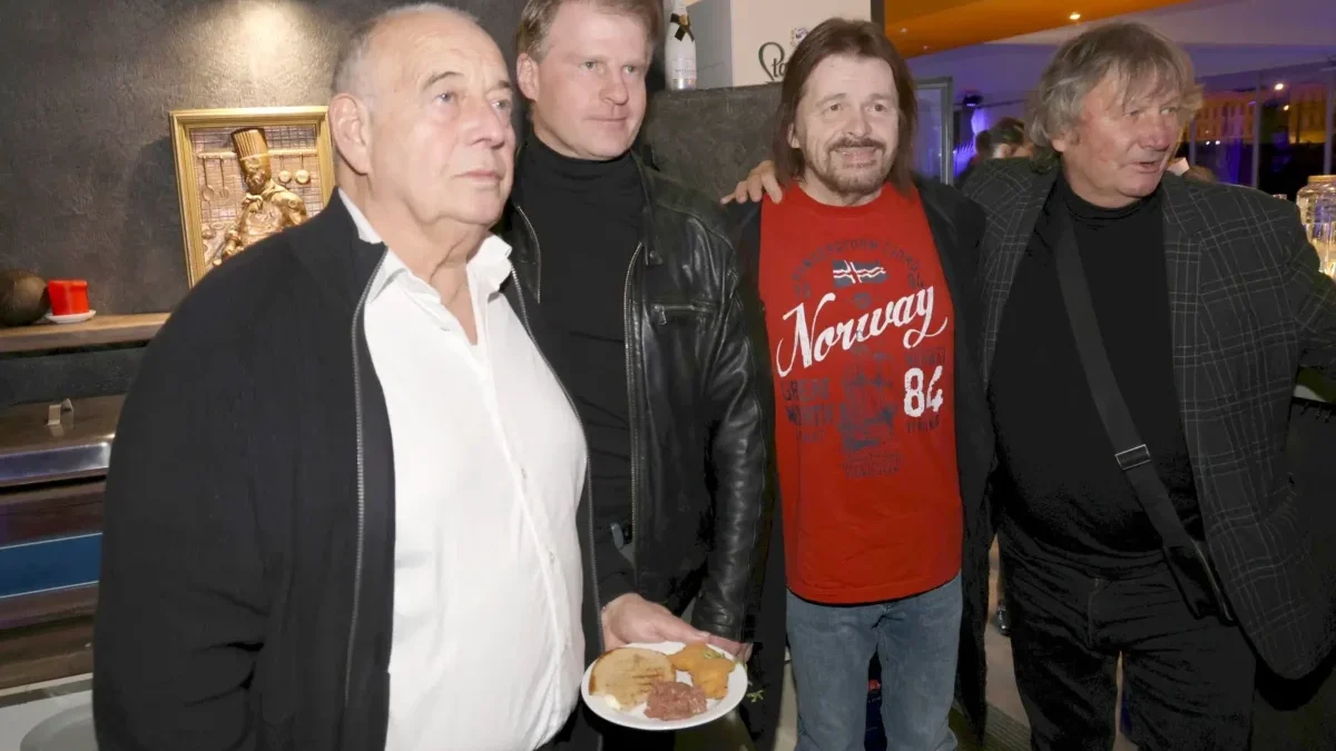Four men at a casual gathering; man in foreground holds a plate with meat and bread while others smile in a kitchen/bar setting behind them.