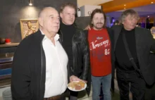 Four men at a casual gathering; man in foreground holds a plate with meat and bread while others smile in a kitchen/bar setting behind them.