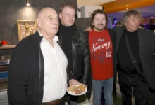 Four men at a casual gathering; man in foreground holds a plate with meat and bread while others smile in a kitchen/bar setting behind them.