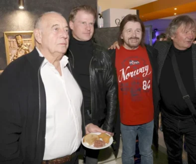 Four men at a casual gathering; man in foreground holds a plate with meat and bread while others smile in a kitchen/bar setting behind them.