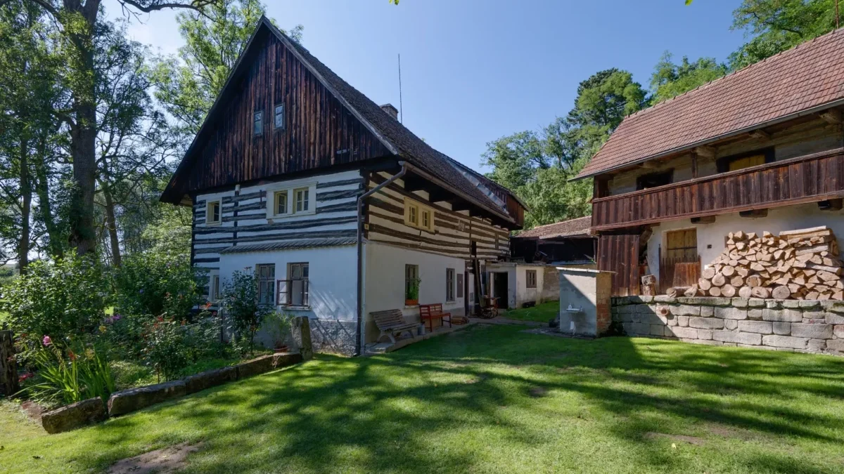 Traditional timber-framed house with white plaster, dark wooden upper level, next to a wooden barn with stacked firewood in a sunny garden.