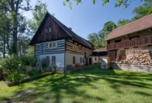 Traditional timber-framed house with white plaster, dark wooden upper level, next to a wooden barn with stacked firewood in a sunny garden.