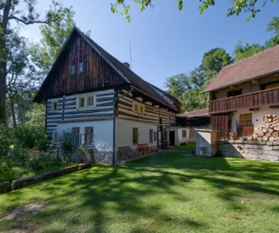 Traditional timber-framed house with white plaster, dark wooden upper level, next to a wooden barn with stacked firewood in a sunny garden.