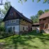 Traditional timber-framed house with white plaster, dark wooden upper level, next to a wooden barn with stacked firewood in a sunny garden.