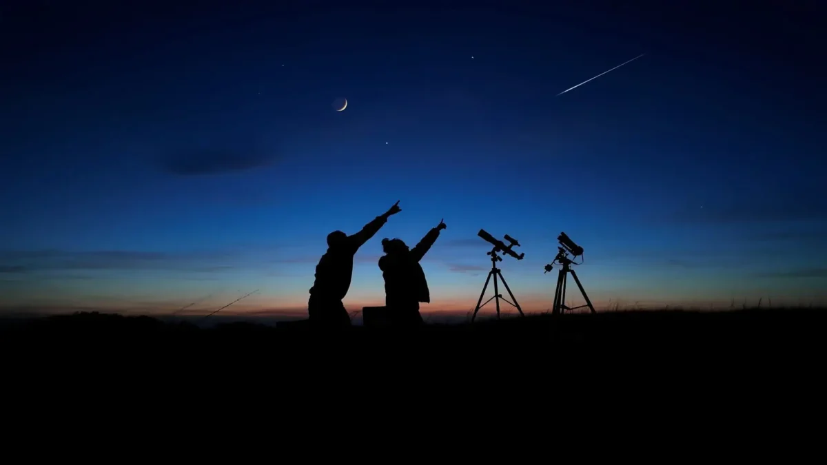 Two people silhouetted at dusk point upward toward a crescent Moon and a shooting star, with two telescopes on tripods nearby.