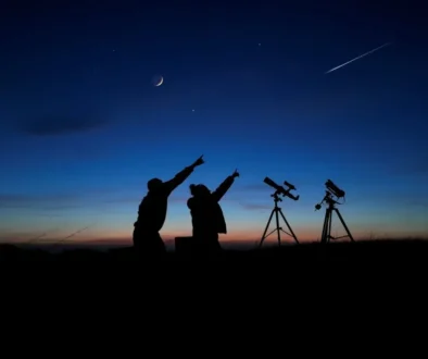 Two people silhouetted at dusk point upward toward a crescent Moon and a shooting star, with two telescopes on tripods nearby.