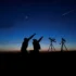 Two people silhouetted at dusk point upward toward a crescent Moon and a shooting star, with two telescopes on tripods nearby.