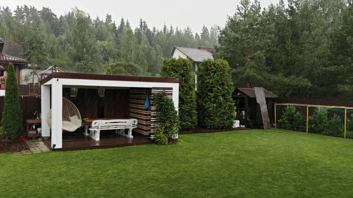 Rainy backyard scene with a white pergola, outdoor seating, and a green lawn under an overcast sky.