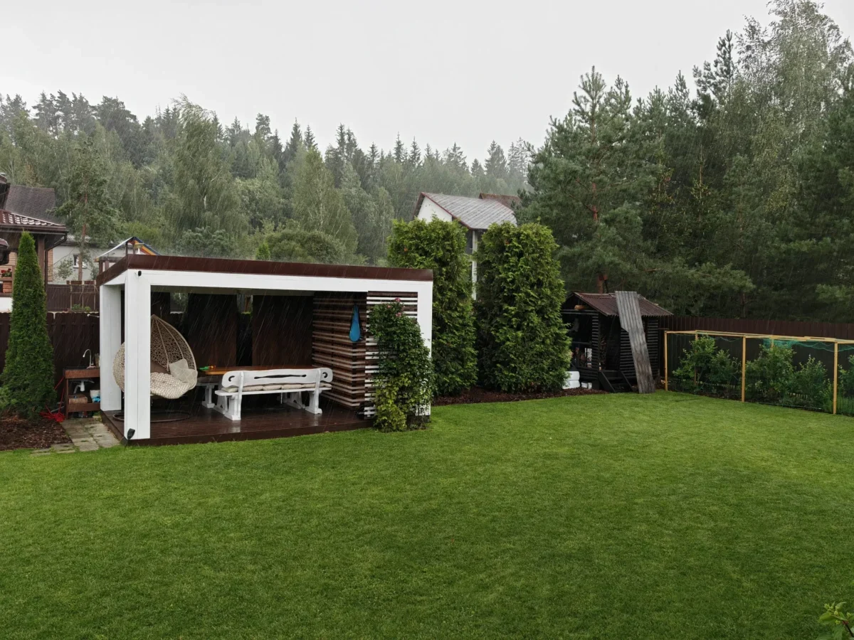Rainy backyard scene with a white pergola, outdoor seating, and a green lawn under an overcast sky.