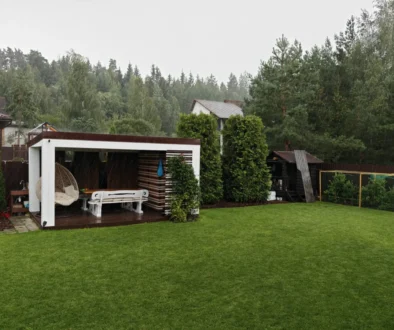 Rainy backyard scene with a white pergola, outdoor seating, and a green lawn under an overcast sky.