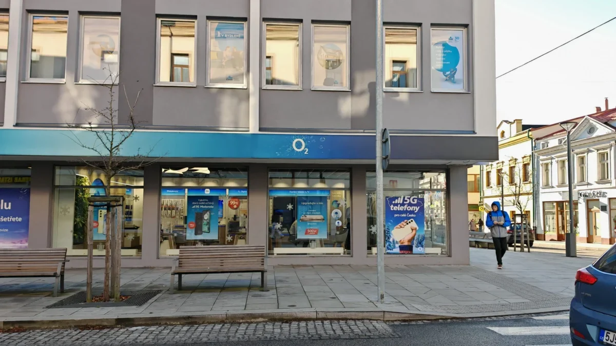 City street storefront with an O2 shop, gray facade, large window posters, and a man in a blue jacket walking nearby.