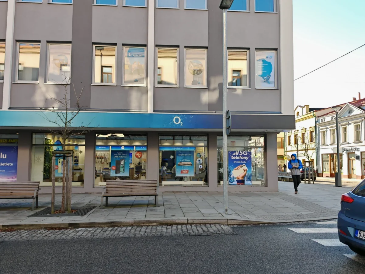 City street storefront with an O2 shop, gray facade, large window posters, and a man in a blue jacket walking nearby.